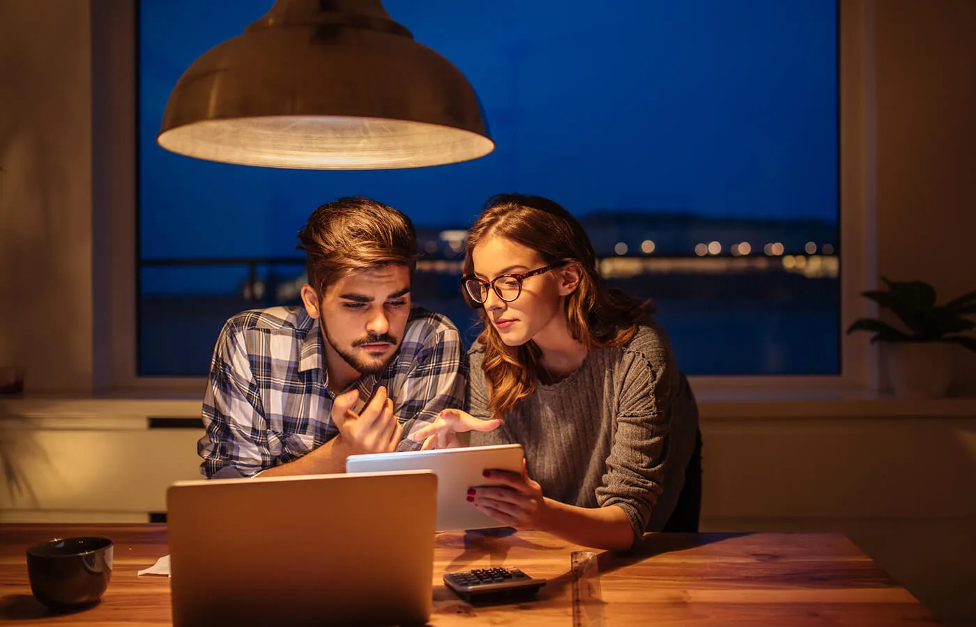 A couple talk with each other as they look at a notebook with a laptop computer on the table.