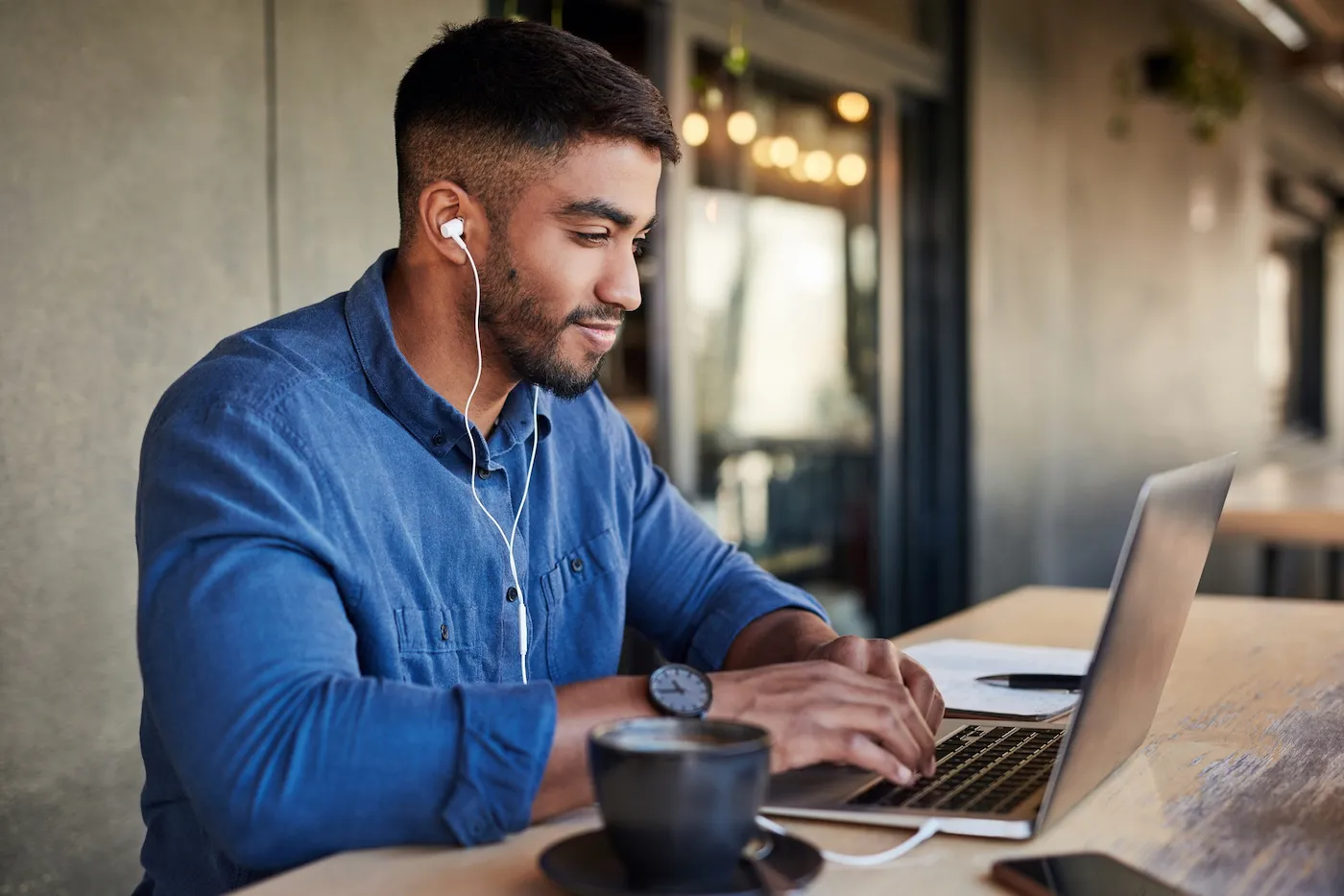 A man sitting at an outdoor table, wearing headphones and typing on his laptop.