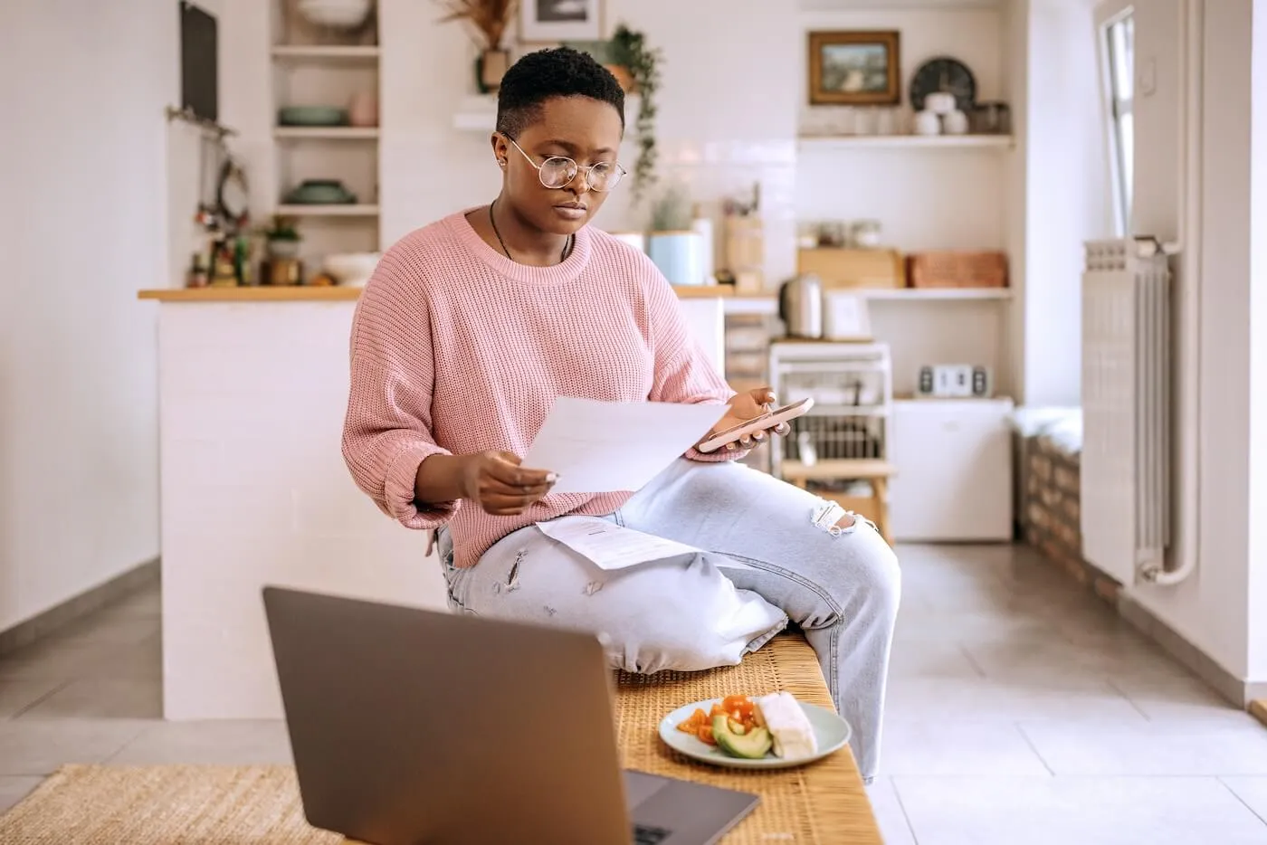 Young woman sitting on a coffee table while reading the printout and holding her smartphone. A laptop and a plate with sandwiches are standing on the coffee table next to her.