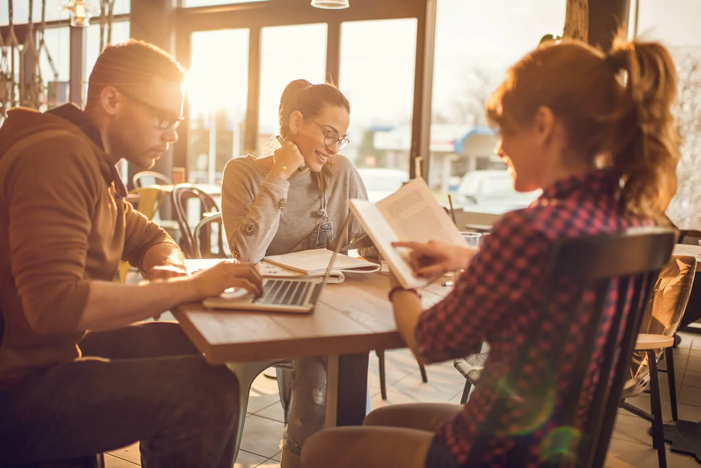 A group of three people are working around a table with one person reading, another person working on their laptop, and another person looking at their phone.