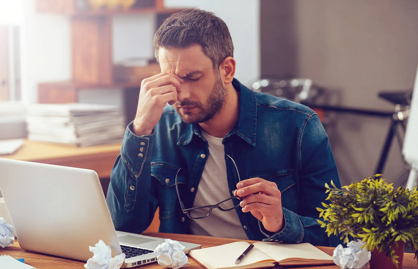 A stressed man pinching the bridge of his nose and holding his glasses as he analyzes his finances.