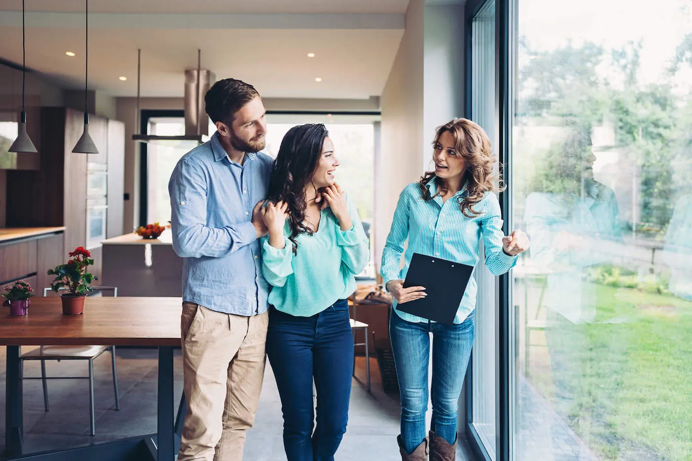 A couple smile and embrace each other as a real estate agent wearing a light blue shirt shows them the backyard of a home.