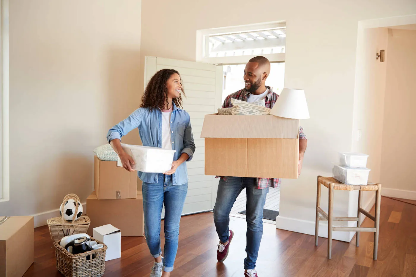 A couple smiles at each other as they hold moving boxes into their new home.