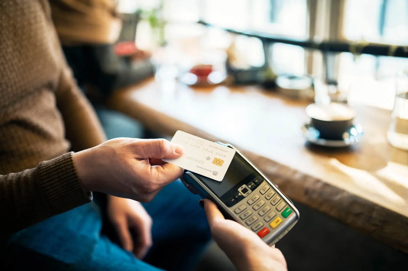 A person wearing a brown sweater taps their credit card on a card reader inside of a coffee shop.