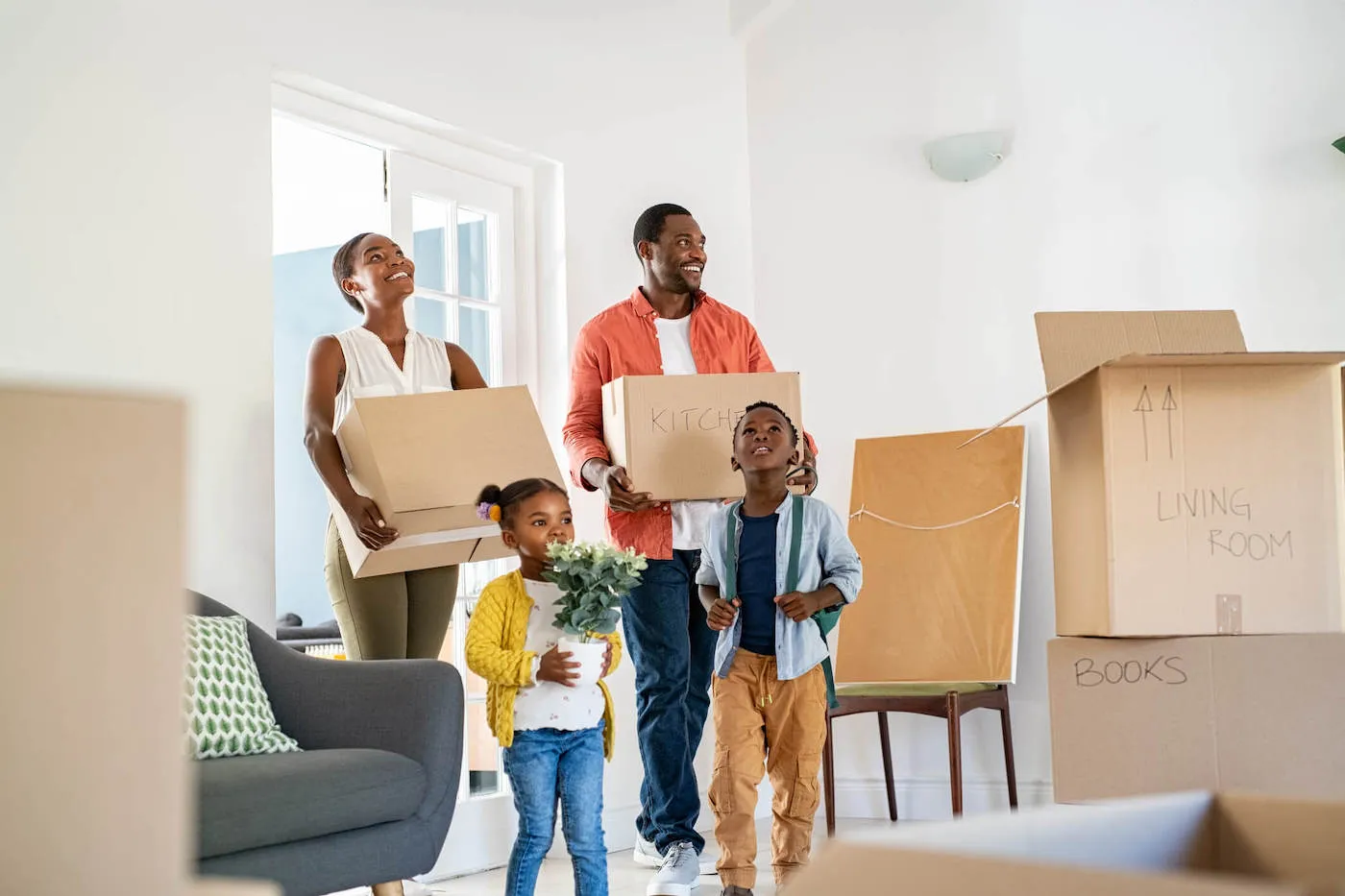 A family of four smiles while carrying boxes into their new home.