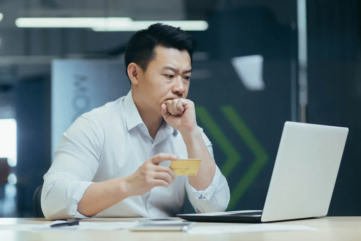 A man with his hand to his face and his credit card in his other hand. frowns while looking at his computer screen.