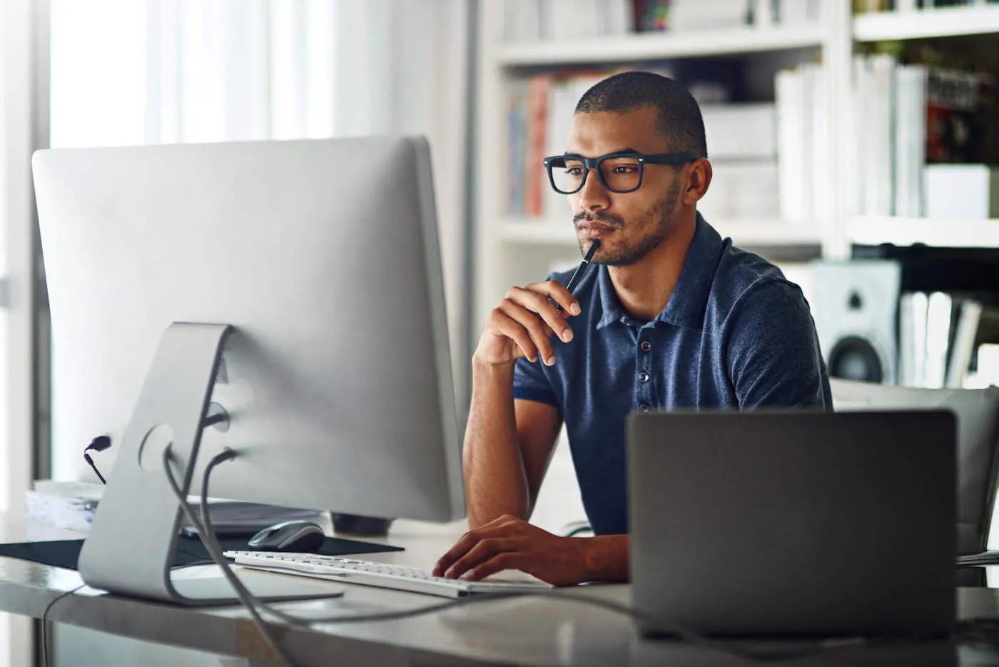 A man wearing a blue shirt and glasses looks at his computer screen as he holds a pen at his work desk.