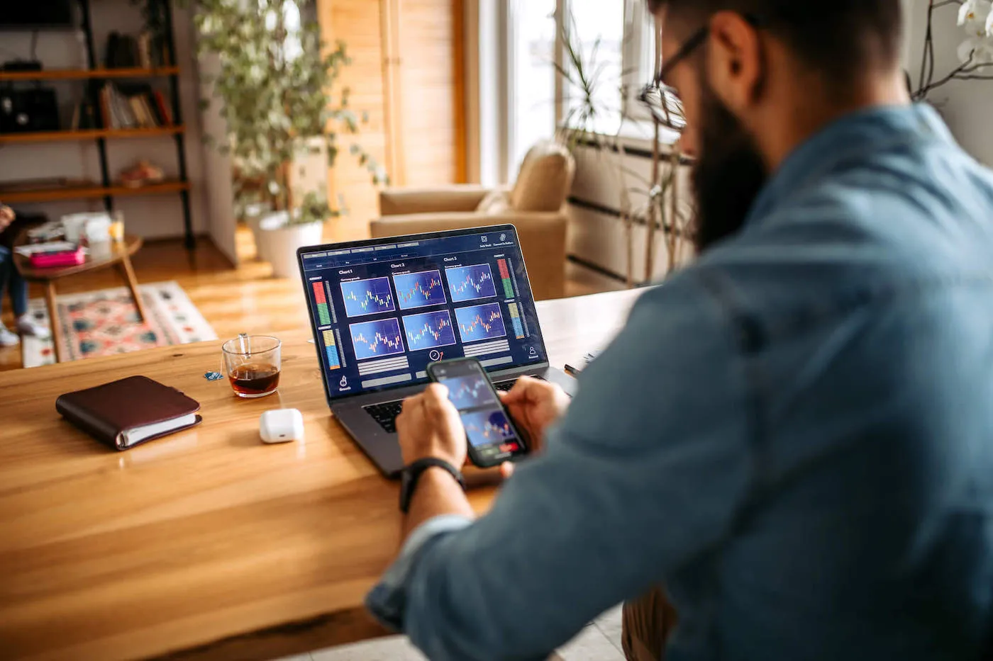 A man wearing a blue shirt looks at stock charts on his phone and computer at home.