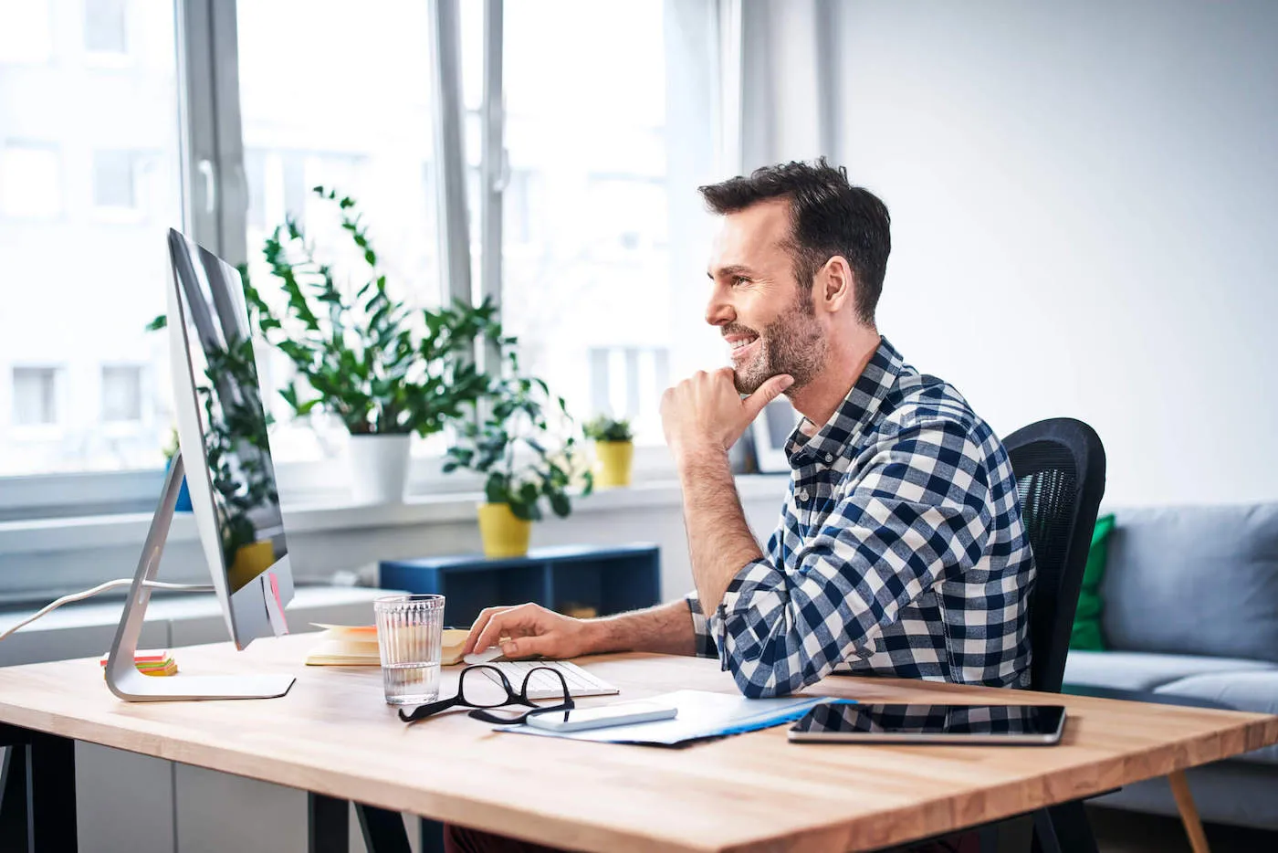 A man wearing a flannel shirt smiles at his computer screen while sitting down at his desk.