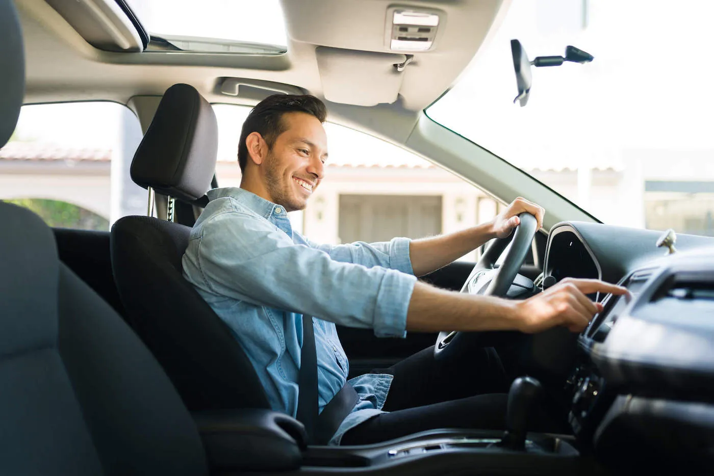 A man wearing a blue shirt smiles as he touches his radio while behind the wheel of his car.