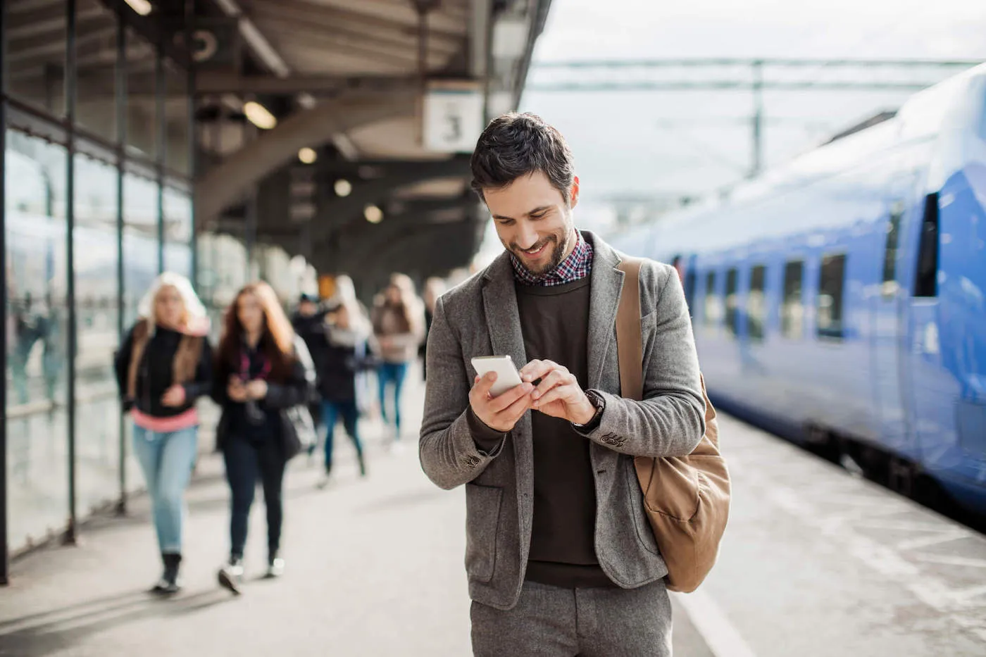 A man wearing a gray suit is smiling at his phone after getting off a blue train at the train station.