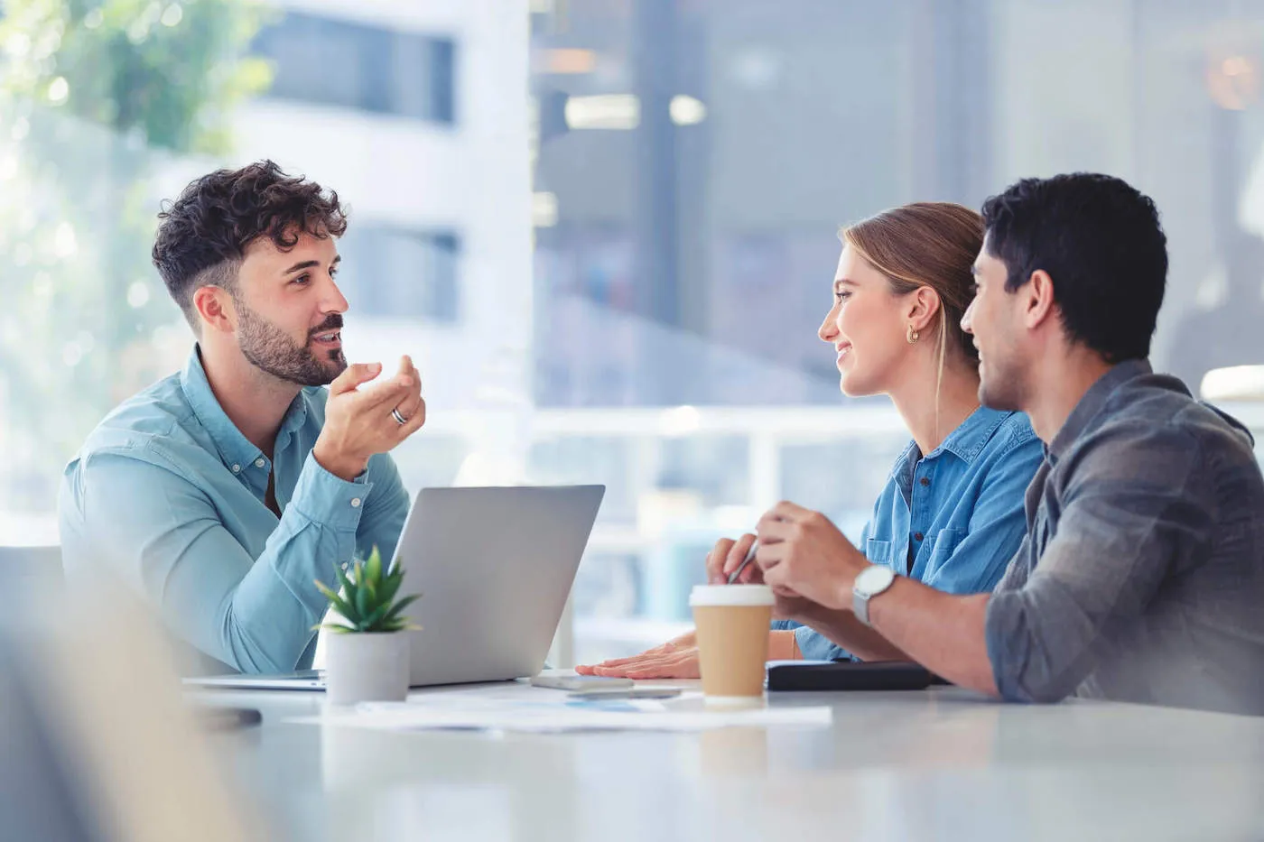 A man wearing a blue shirt talks to a couple from across the table while his laptop is in front of him.