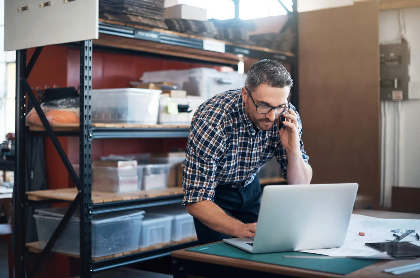 A man wearing a flannel shirt uses his laptop while talking on the phone with materials in the background.