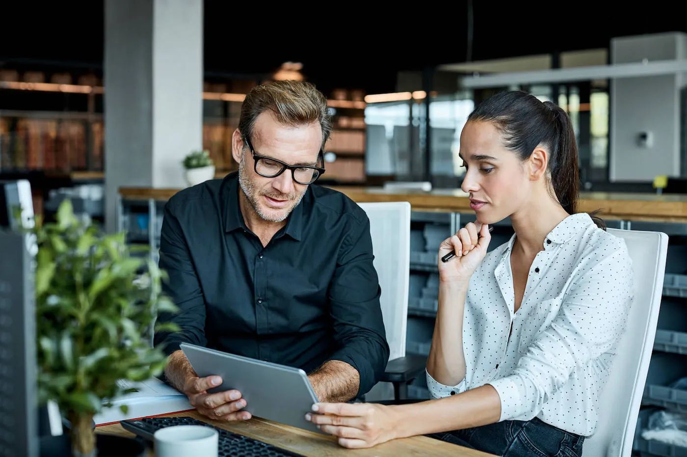 A man wearing a black button-up shirt is showing his female client a tablet at his office.