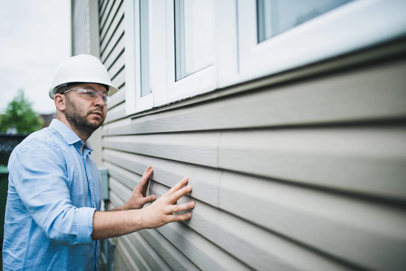 A man wearing a blue shirt, hard hat, and safety glasses inspects the walls of a home.