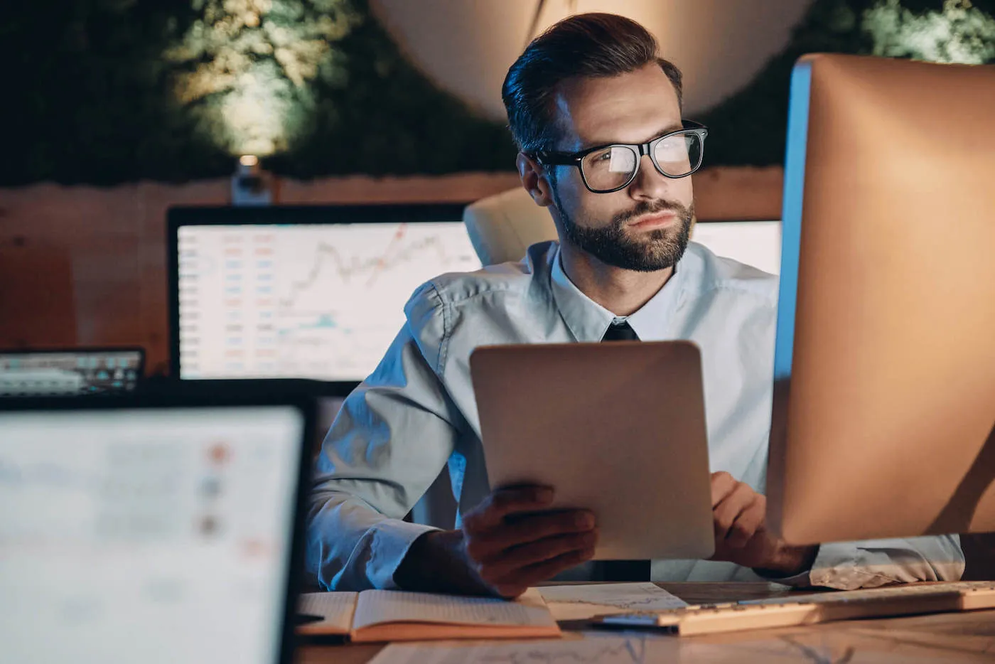 A man wearing black glasses and a shirt with tie is looking at his computer screen while holding a document.