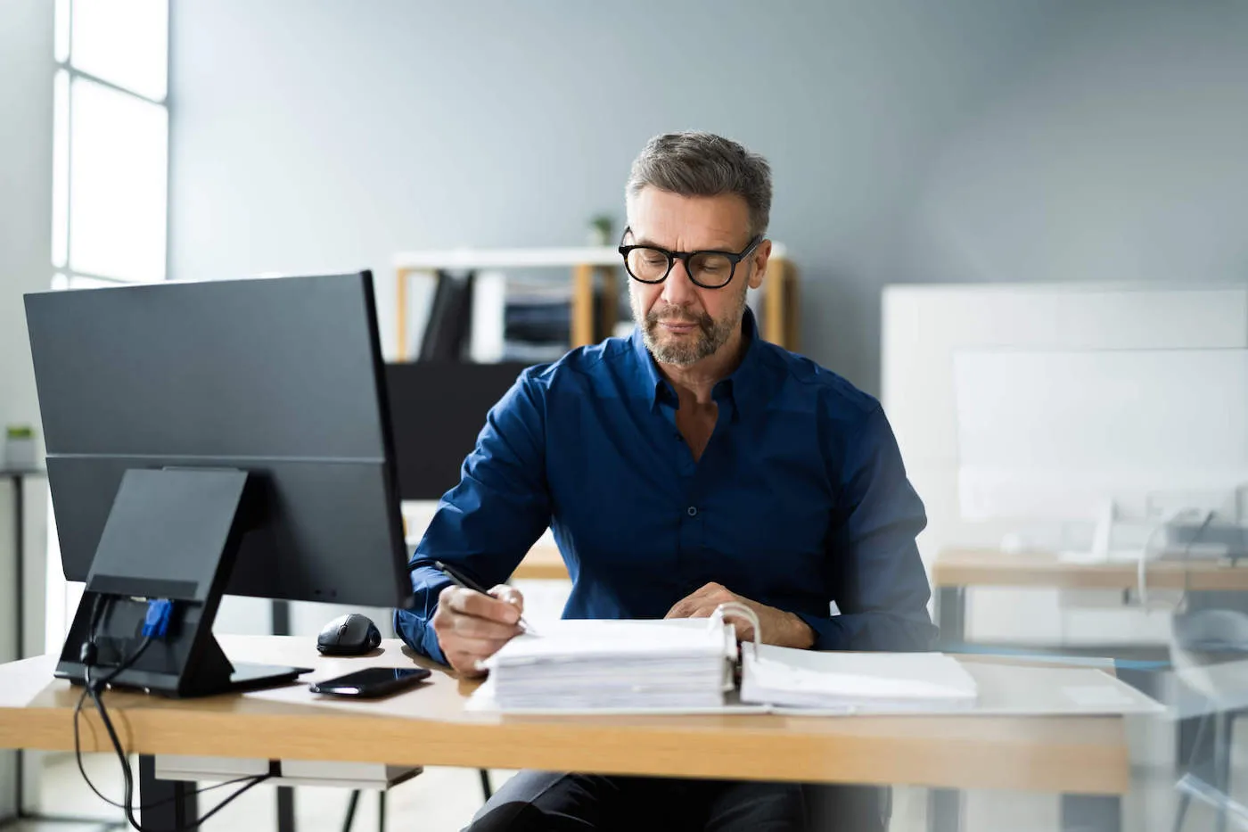 A man wearing glasses looks at documents while in front of his work computer.