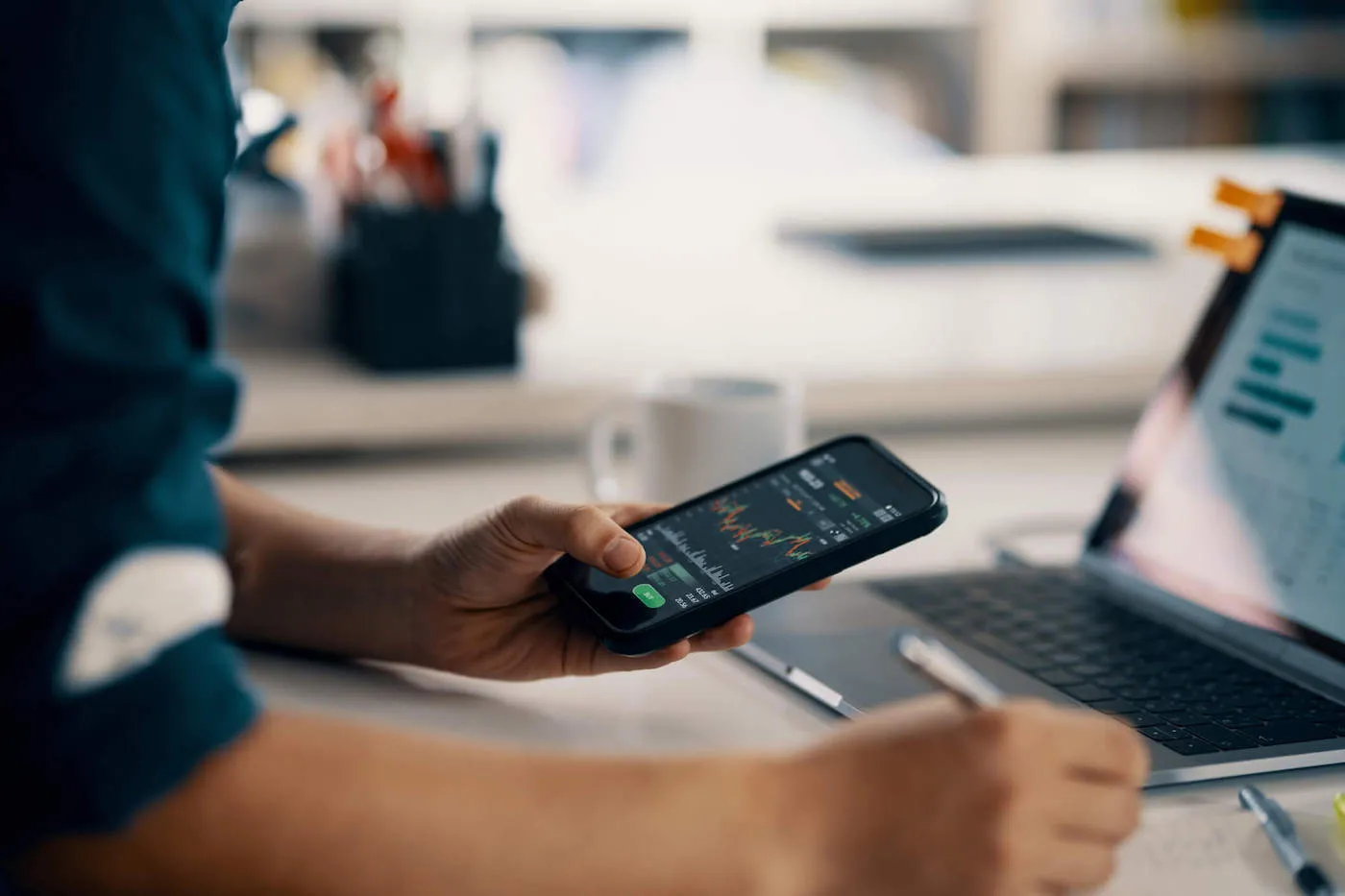 A person checks a stock graph on their phone while holding a pen and writing on a notebook.