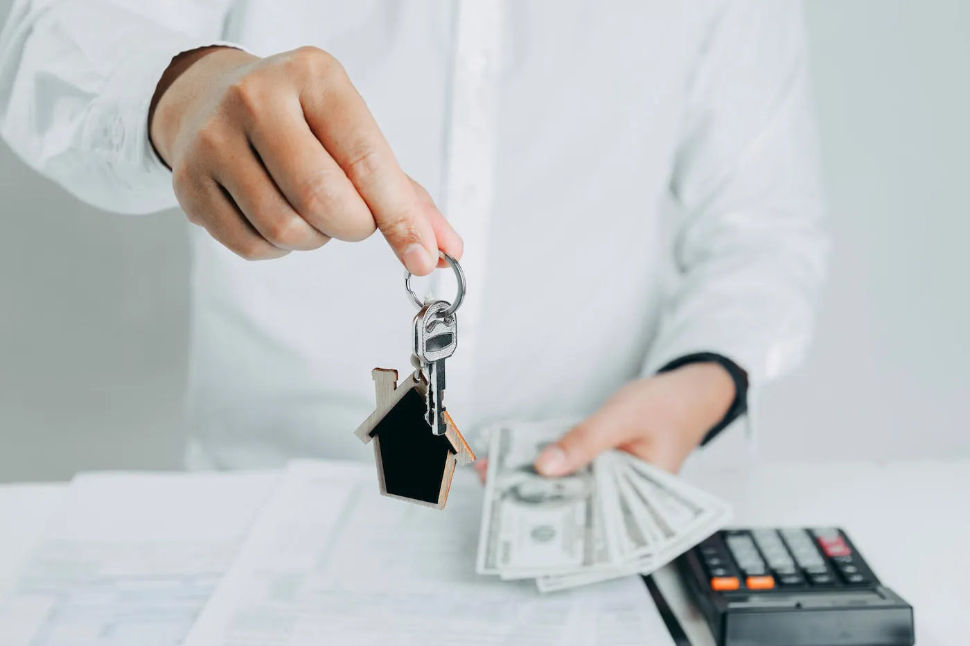 A person wearing a white shirt is holding up house keys on one hand while holding cash in the other.