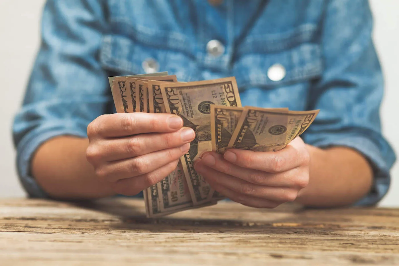 A person wearing a blue shirt is counting fifty dollar bills on the table.