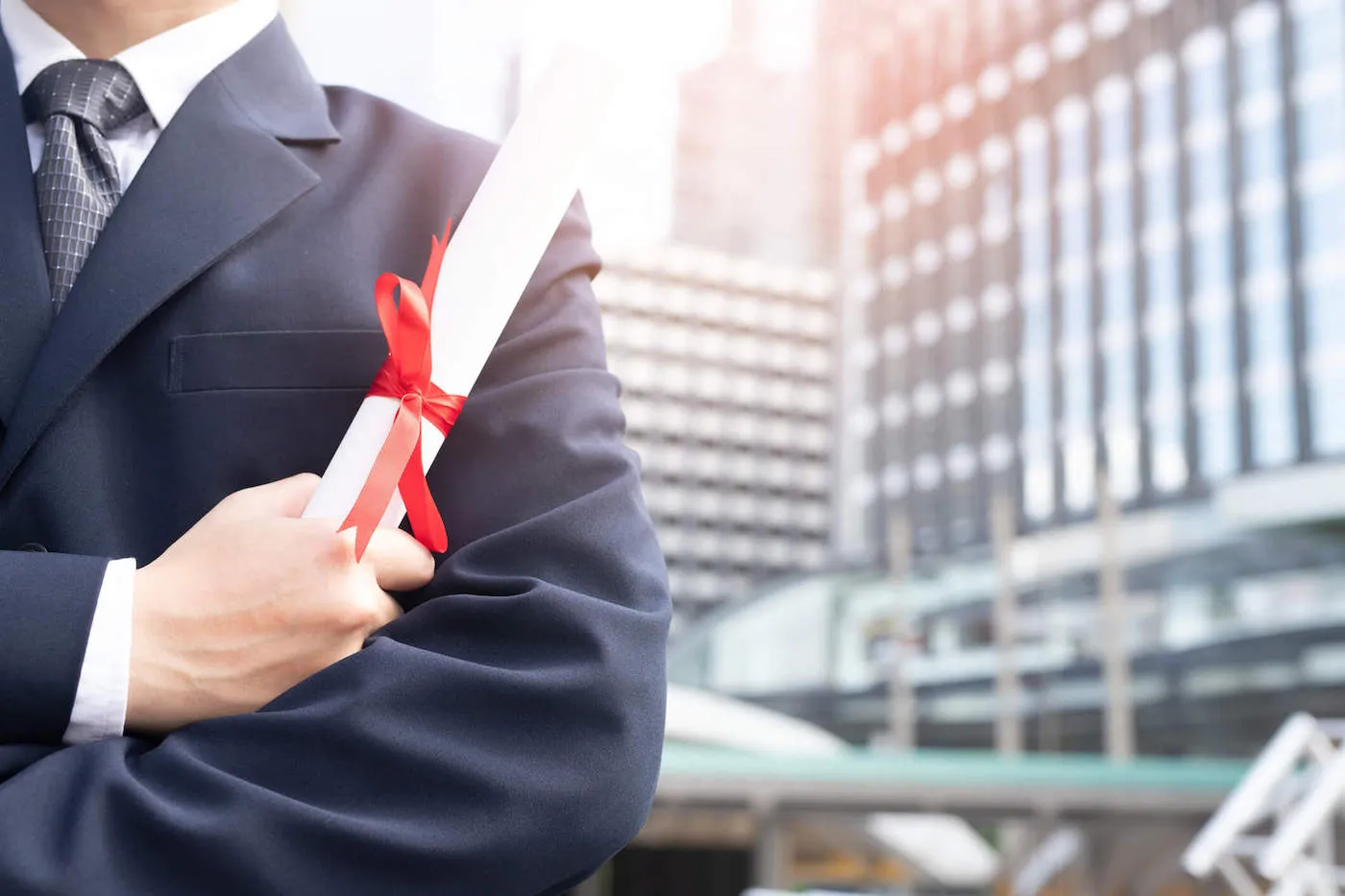 A person wearing a black suit holds their diploma with skyscrapers in the background.