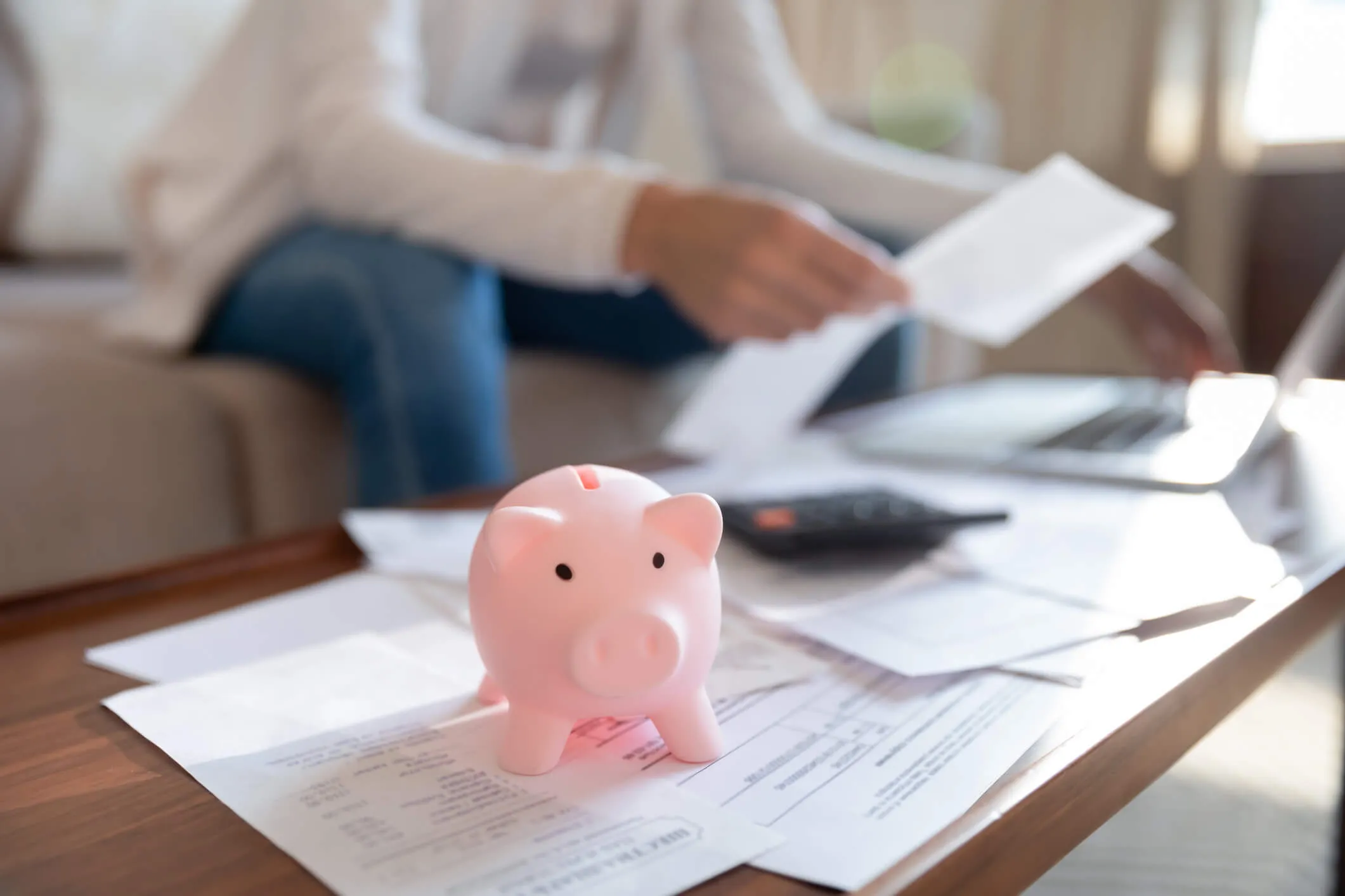 A pink piggy bank sits on top of documents on the living room table while someone type on a laptop in the background.