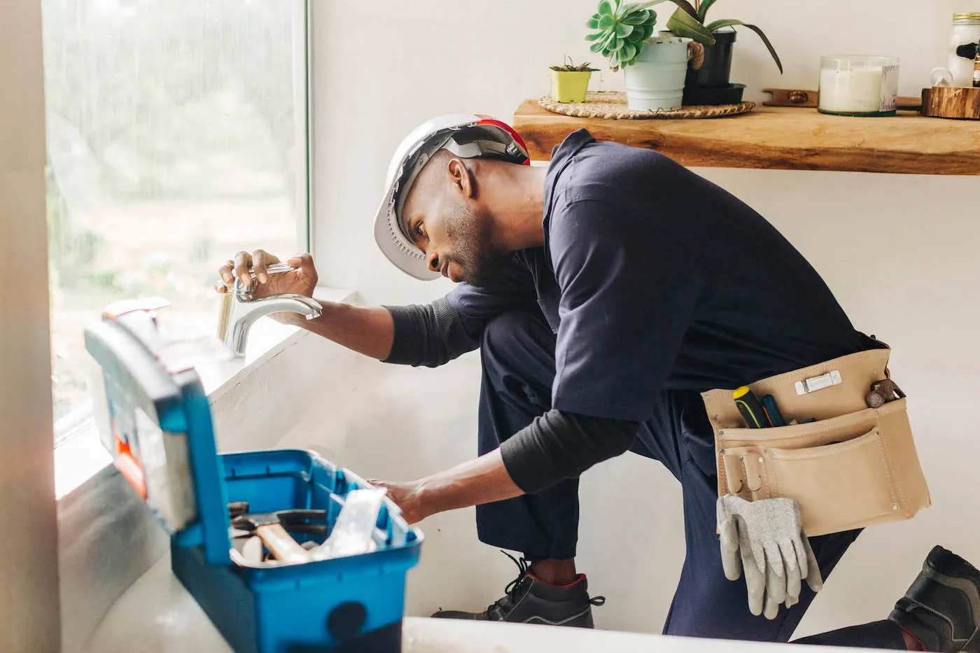 A plumber wearing a blue uniform looks at a bathroom faucet with his tools to the side.