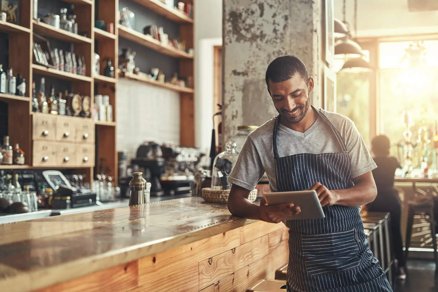 A small business owner wearing a blue apron smiles while looking at a tablet.