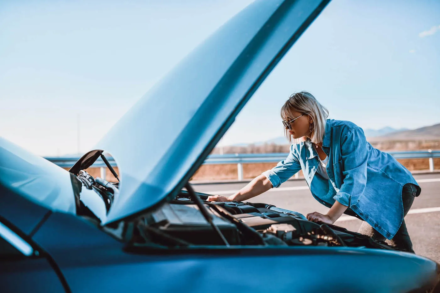 A woman wearing a blue jacket and sunglasses checks the engine of her card by the side of the road.