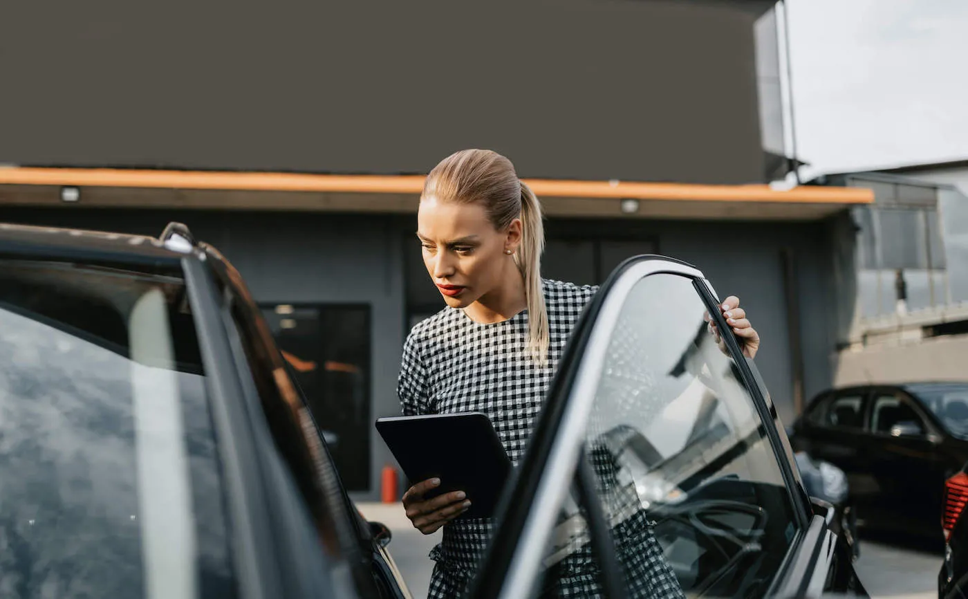 A woman wearing a tablet check out a car that is parked outside.