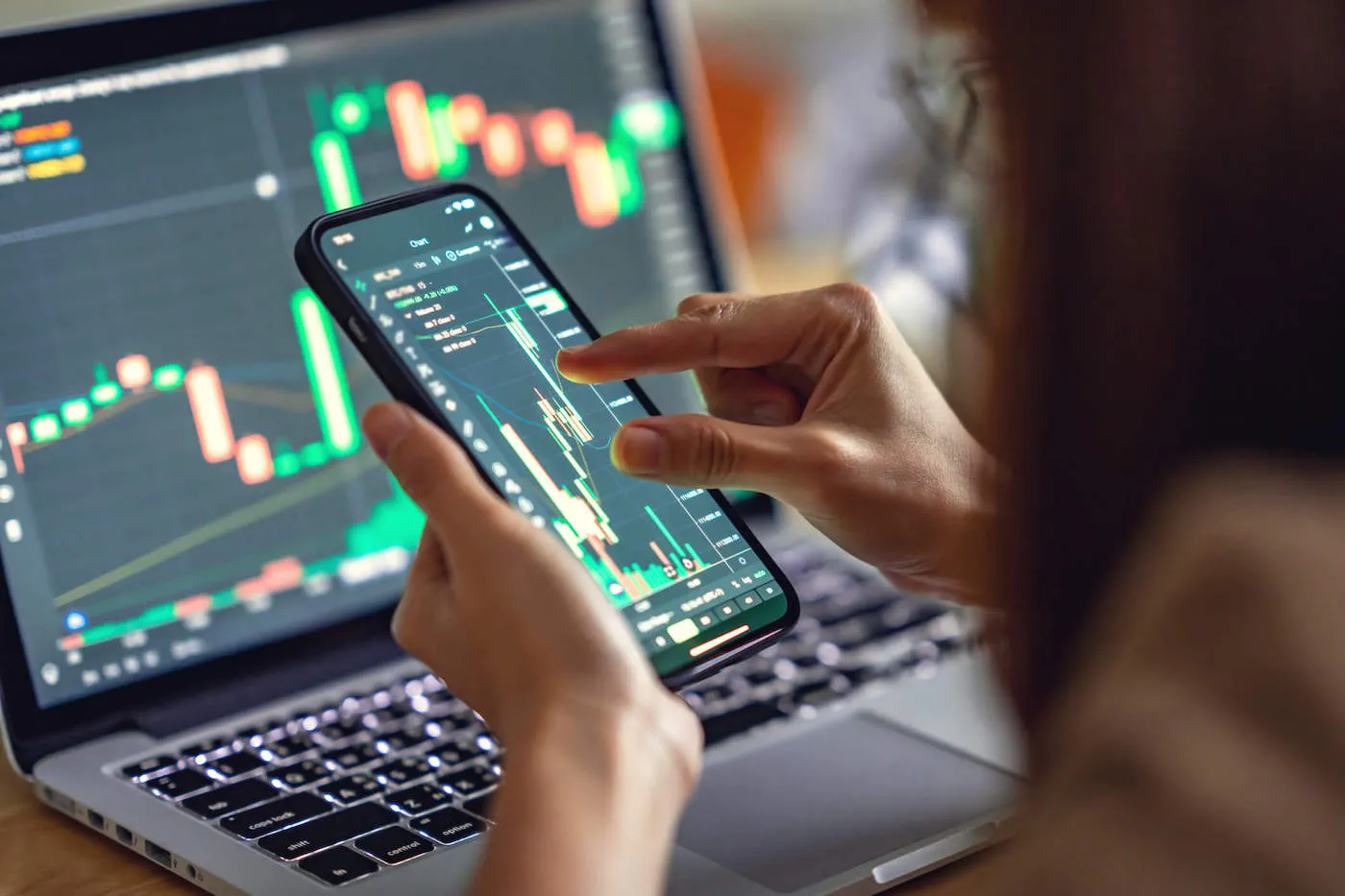 A woman is holding her phone while looking at stock charts on her phone and laptop.