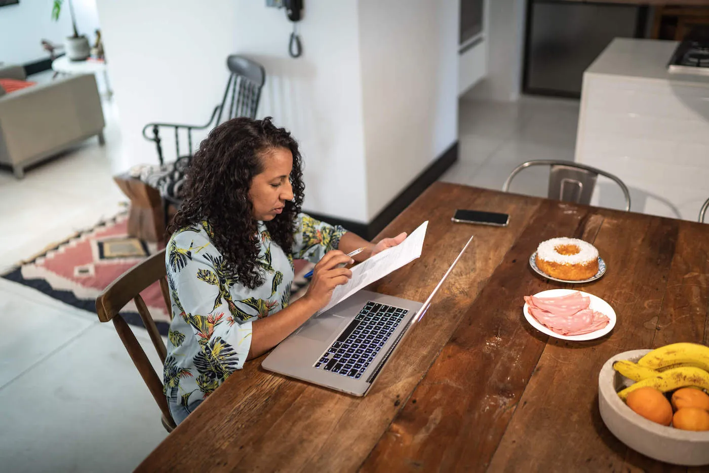 A woman looks at a document next to her laptop while sitting at the kitchen table.