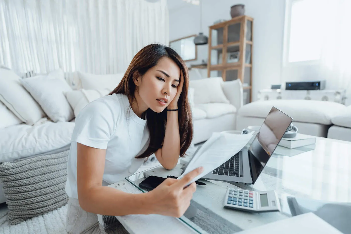 A woman leans on her hand while looking at documents with her laptop and calculator at the living room table.