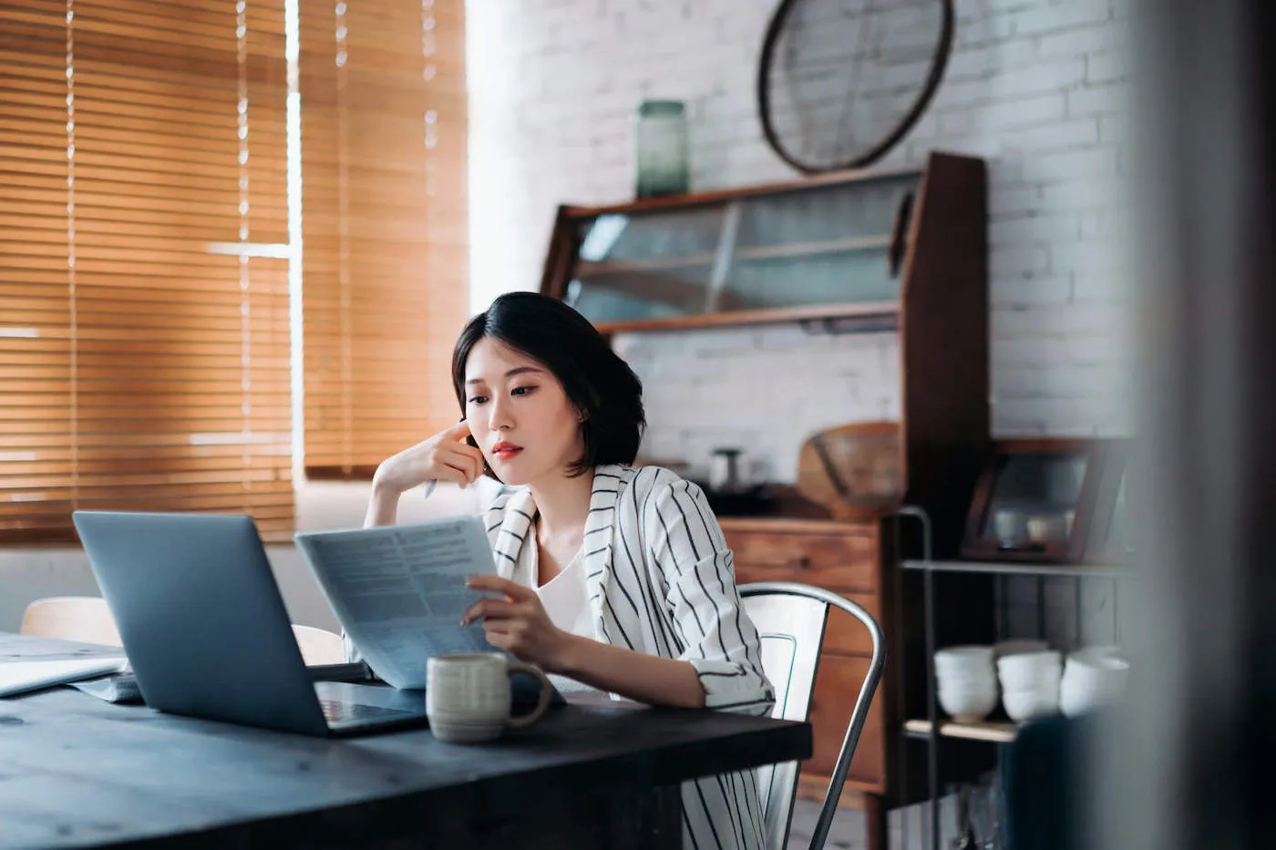 A woman sitting down looks at a document while her laptop sits on the table.