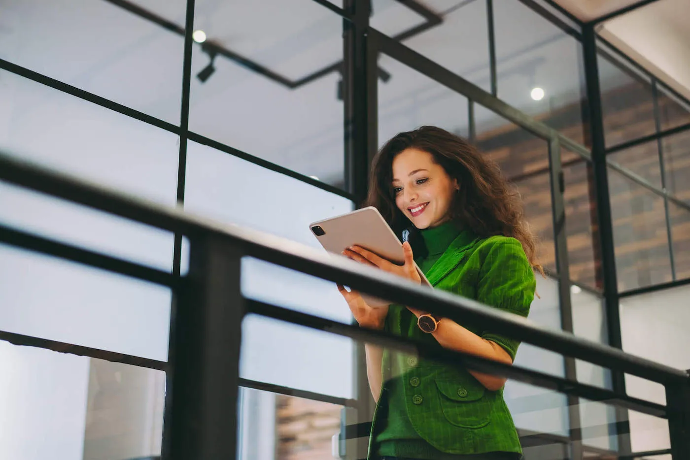 A woman wearing a green suit and turtleneck smiles at a tablet screen she is holding at the work office.