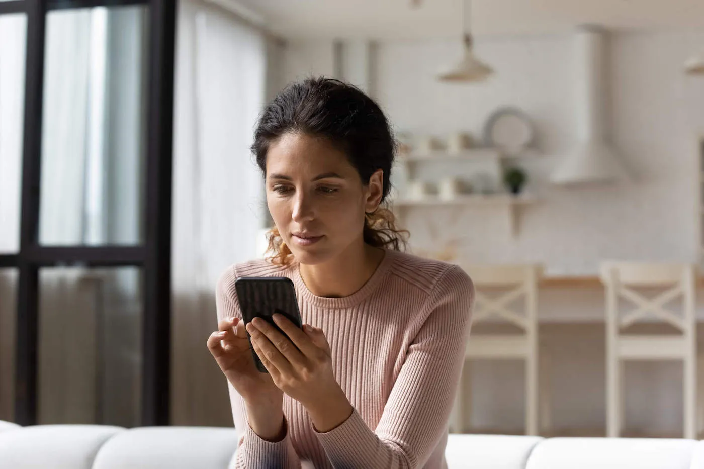 A woman wearing a pink sweater uses her phone while sitting on the couch at home.