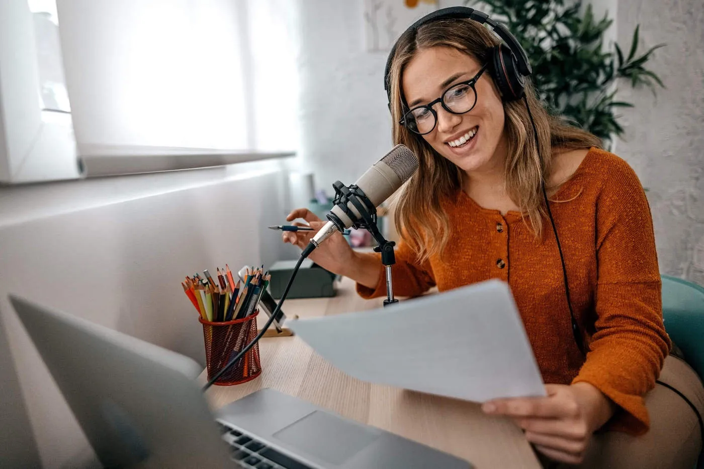 A woman wearing headphones smiles as she talks into a microphone while holding a document and pen in her hands.