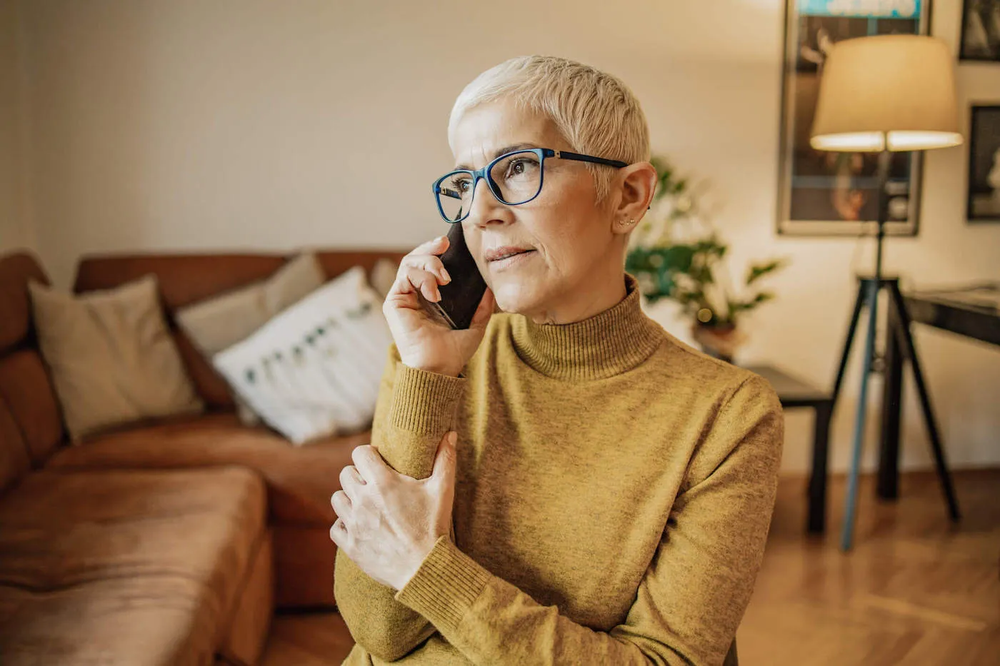 A women wearing a yellow sweater and black glasses is on the phone at her living room.