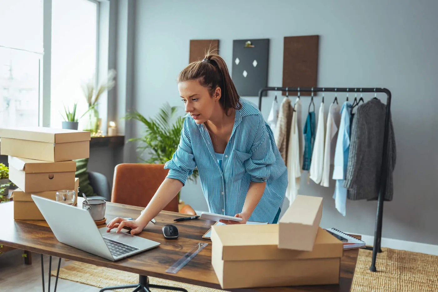 A young business owner looks at her laptop next to shoes boxes piled up on her work desk.