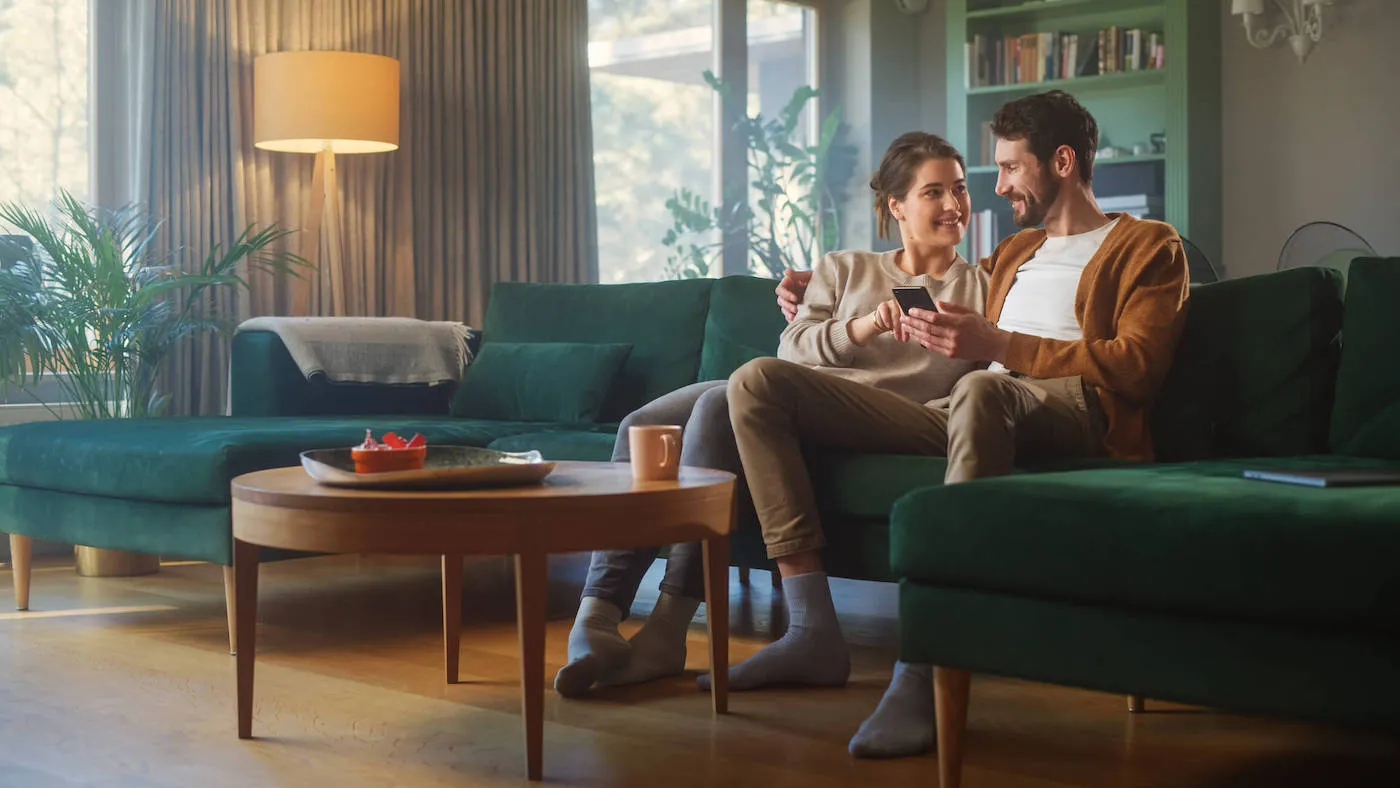 A young couple smile at each other while holding a phone and sitting on a green couch.