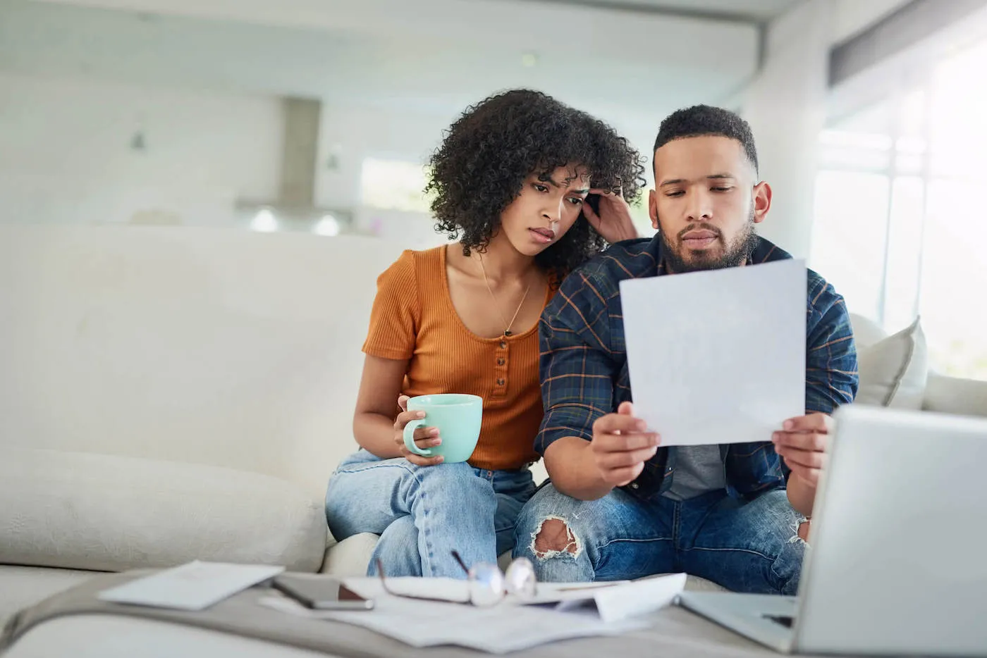 A young couple sits on their couch as they look at a document together while their laptop is on the table.