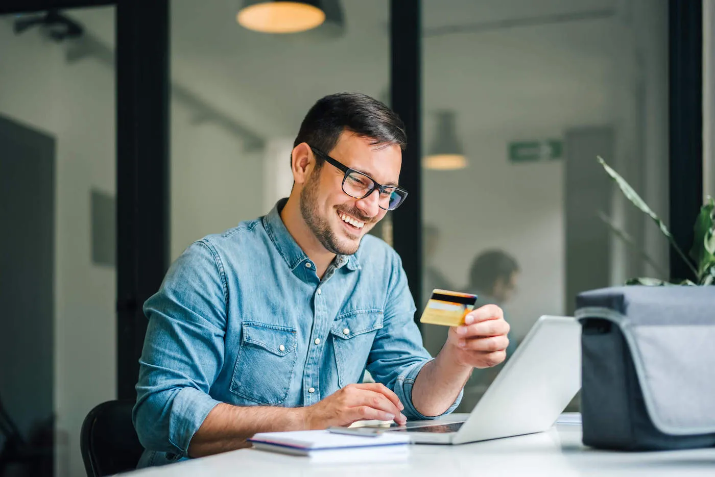 A young man wearing holding his credit card smiles while looking at his laptop in his office.