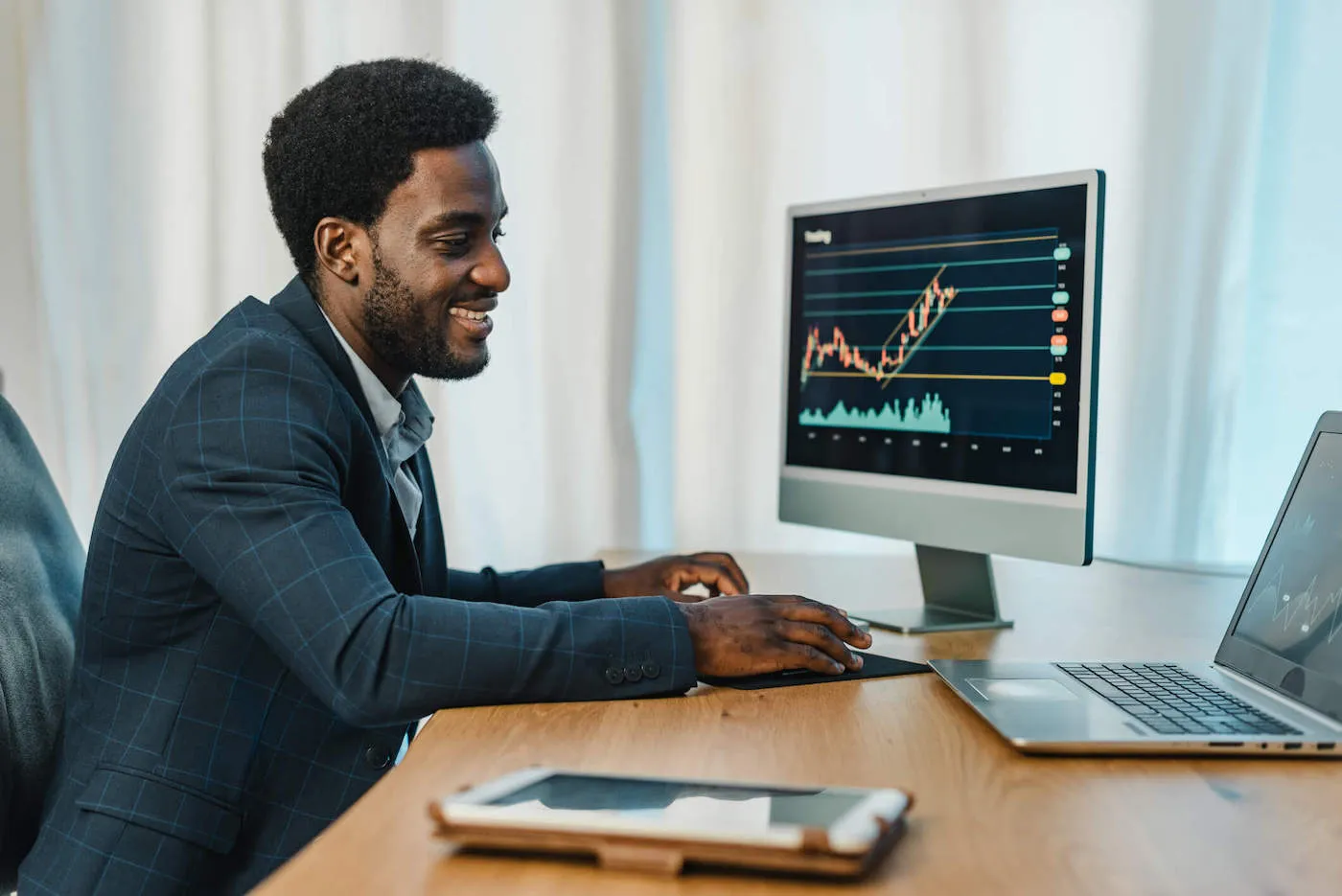 A young man wearing a blue suit smiles while looking at computer screens showing stock charts.