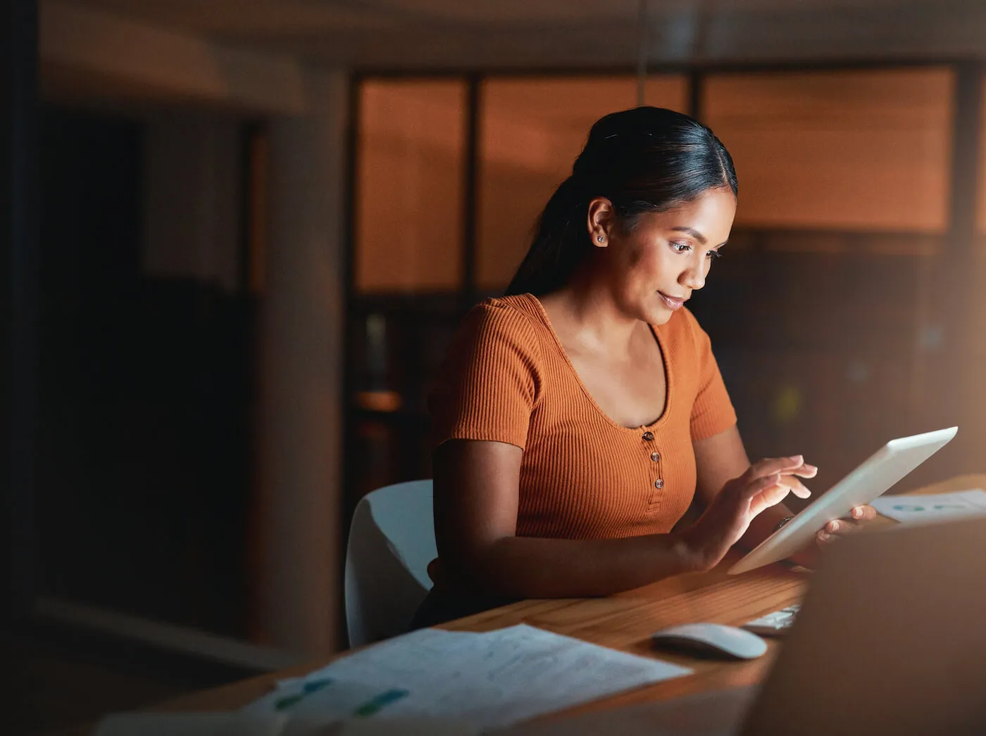 A young woman wearing an orange shirt is using a tablet while working at her desk.