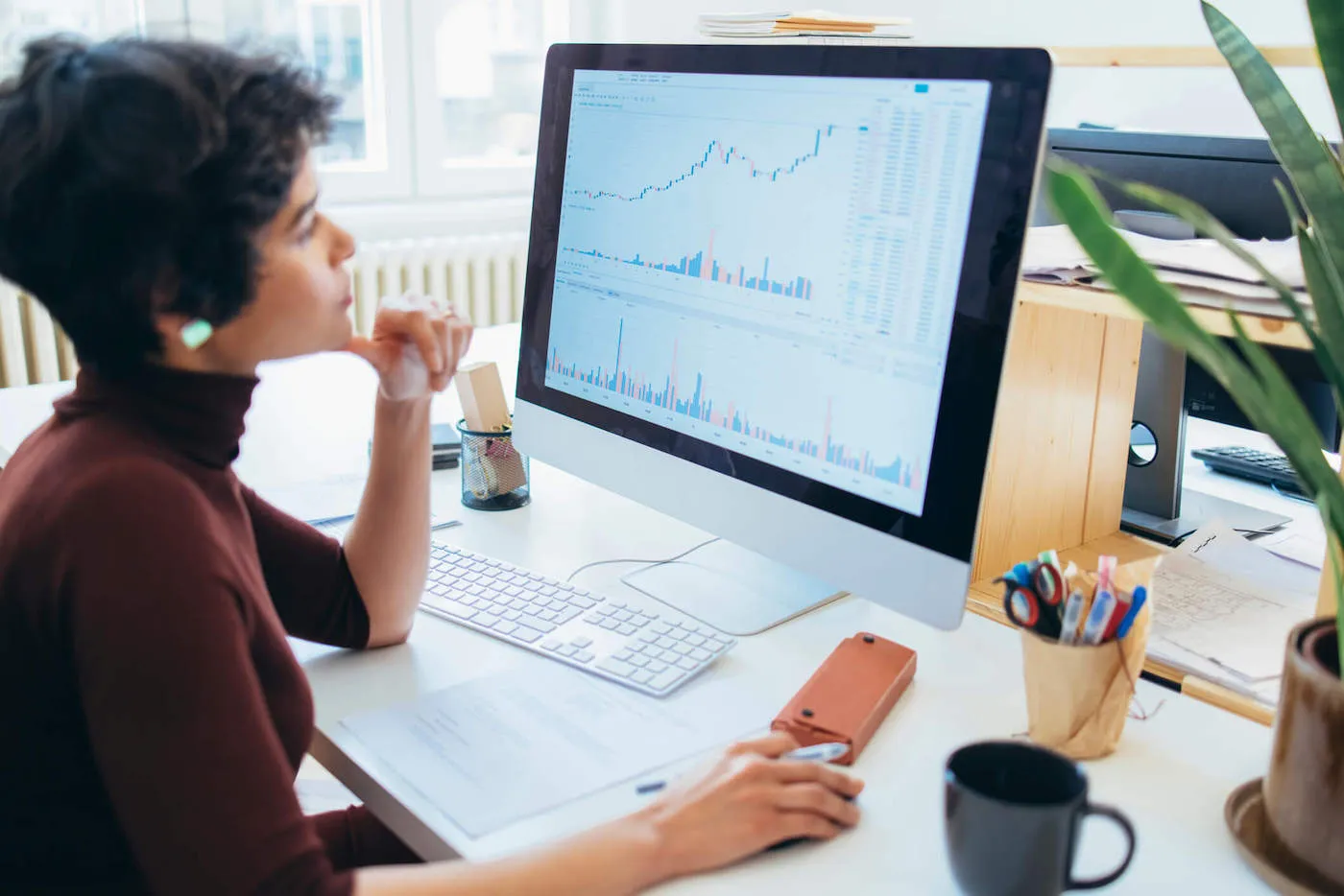 A young woman wearing a red sweater looks at a stock chart on her computer.