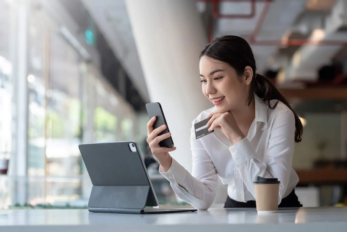 A young woman wearing a white shirt smiles at her phone as she holds her credit card.