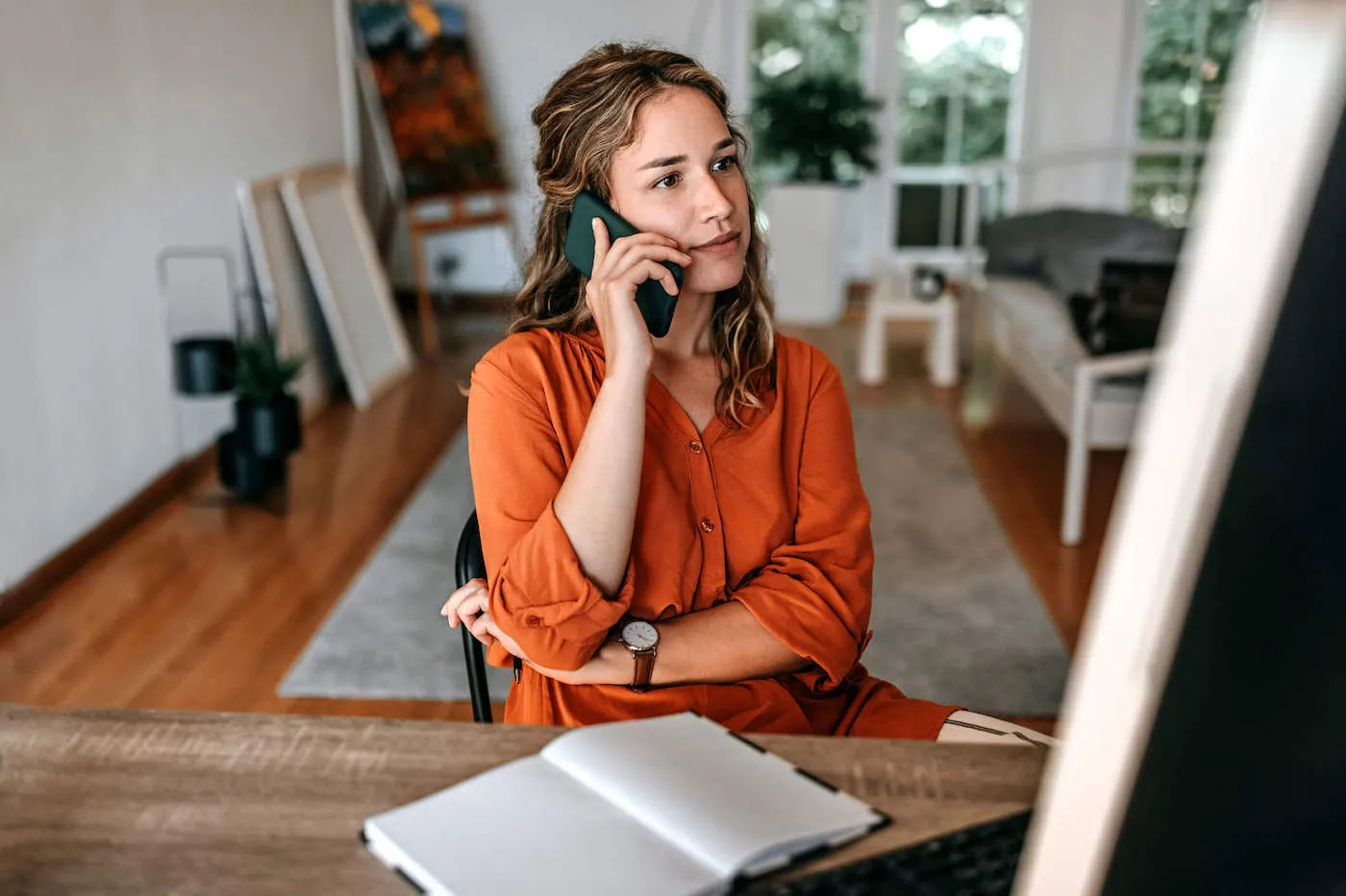 A young woman wearing an orange shirt talks on the phone while her notebook is on the table in front of her.