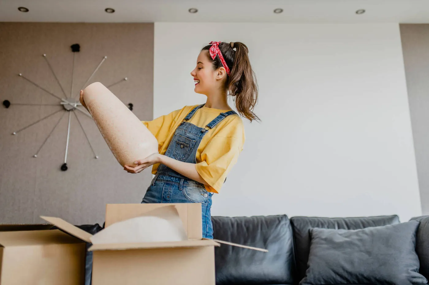 A young woman wearing a yellow shirt smiles at a vase she just opened from a box in her living room.