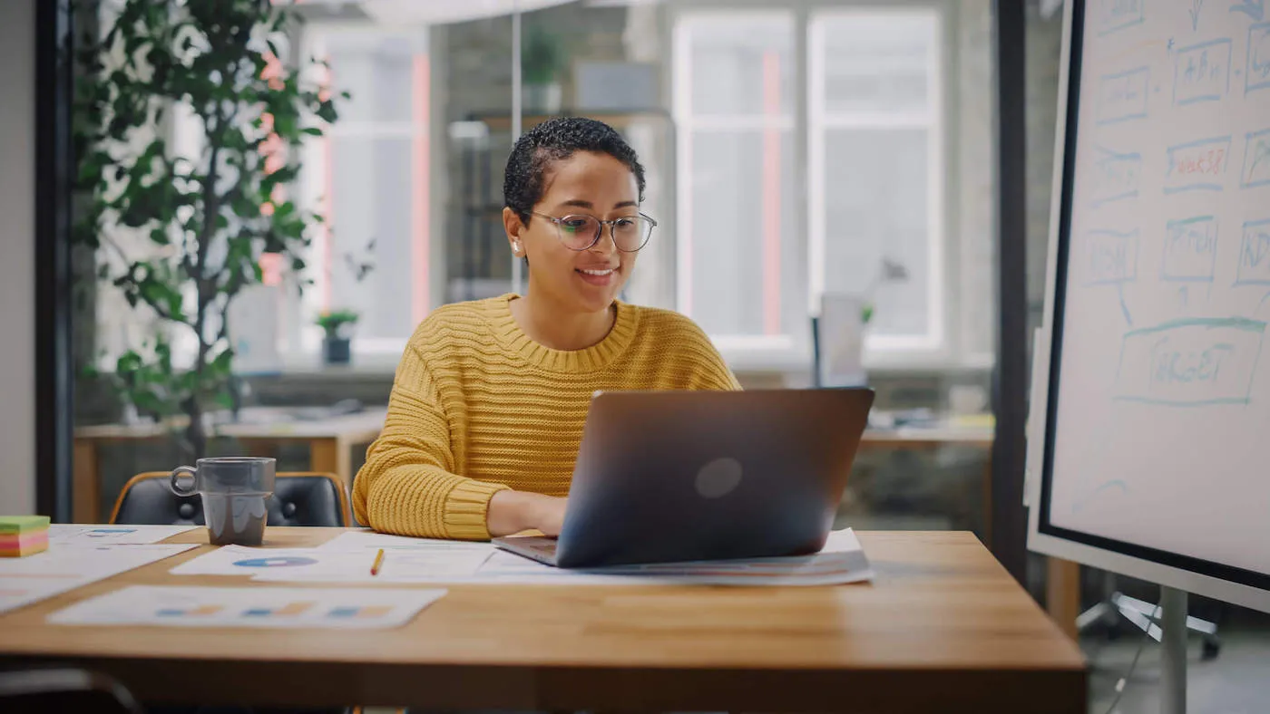 A young woman wearing a yellow sweater and glasses smiles as she uses her laptop.