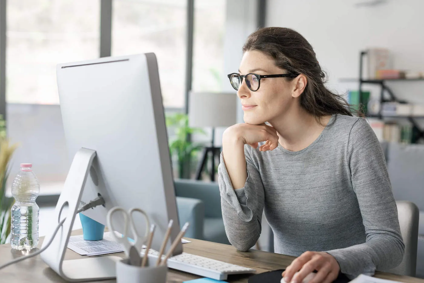 A young woman wearing a gray shirt and glasses looks at her computer screen from her office.